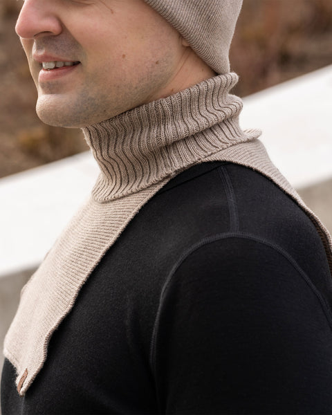 A close-up profile shot focusing on a man's lower face, neck, and chest as he faces left with a slight smile. He is wearing a beige knit neck warmer featuring a ribbed turtleneck collar and a flat front bib over a black long-sleeve shirt. A small brown logo patch is visible on the bottom corner of the bib. The bottom edge of a matching beige knit beanie is visible on his head. The background is a blurred outdoor scene featuring concrete and brown earth tones.
