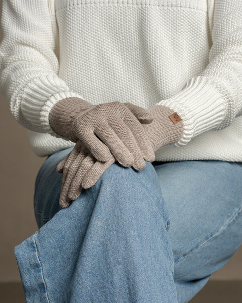 Person wearing creamy beige merino wool knit gloves, hands resting on denim-clad knee, showcasing soft texture and comfortable fit.