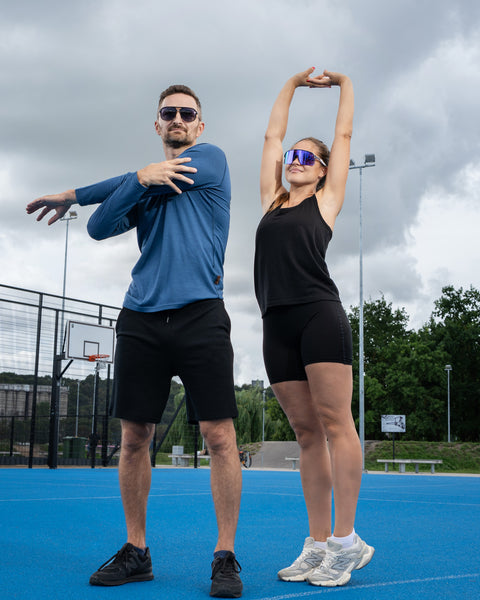 A man and a woman stretching side-by-side on an outdoor blue sports court. The man is wearing a blue long-sleeved shirt and stretching his arm across his chest, while the woman wears black athletic wear and stretches her arms upward.