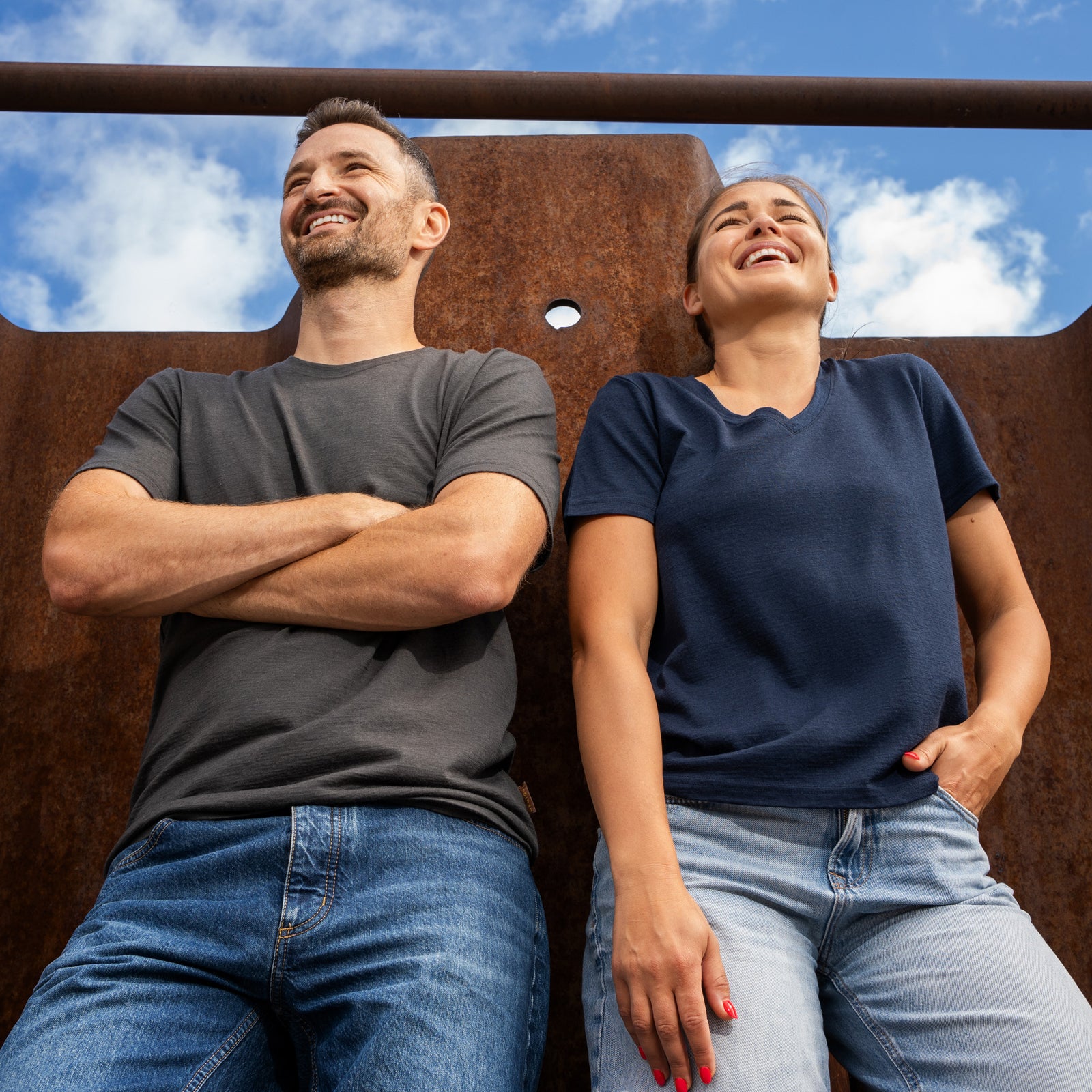 A low-angle shot of a smiling man and a laughing woman leaning back against a large, rusted metal wall under a blue sky with clouds. The man is on the left with his arms crossed, wearing a dark grey short-sleeve t-shirt and blue jeans. The woman is on the right with one hand resting in her pocket, wearing a navy blue short-sleeve v-neck t-shirt and light blue jeans.