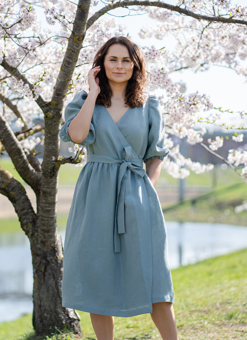 Woman wearing a blue linen midi wrap dress standing outdoors by a pond under blooming cherry blossom trees on a sunny spring day.