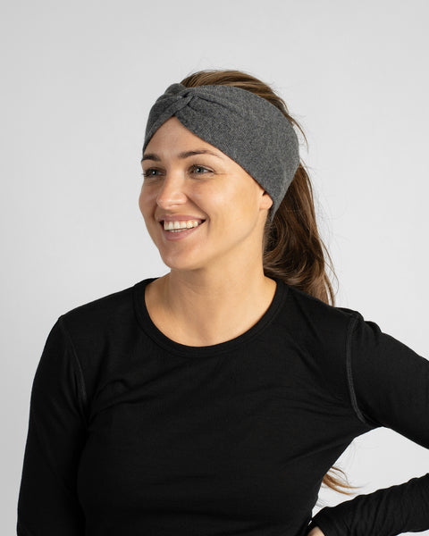 A smiling woman with her hair pulled back into a ponytail, wearing a dark gray or charcoal wide knit headband and a black long-sleeve top.