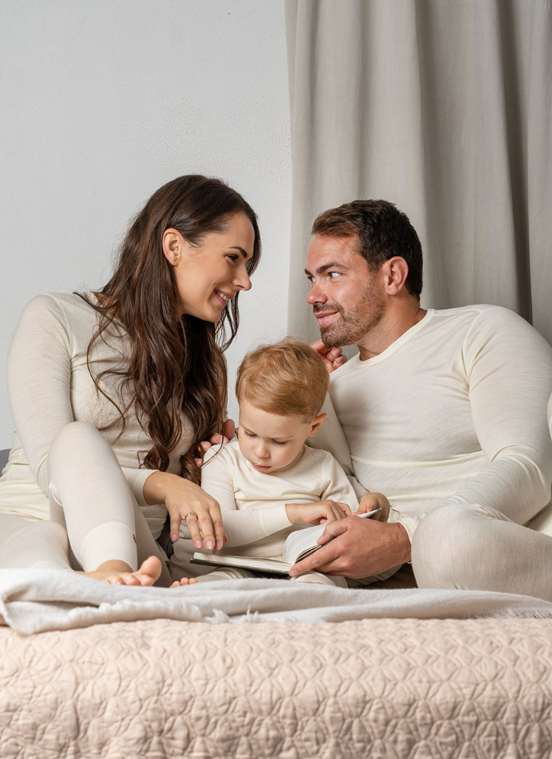 Family of mom, dad and kid sitting on the bed and wearing matching natural merino wool sets