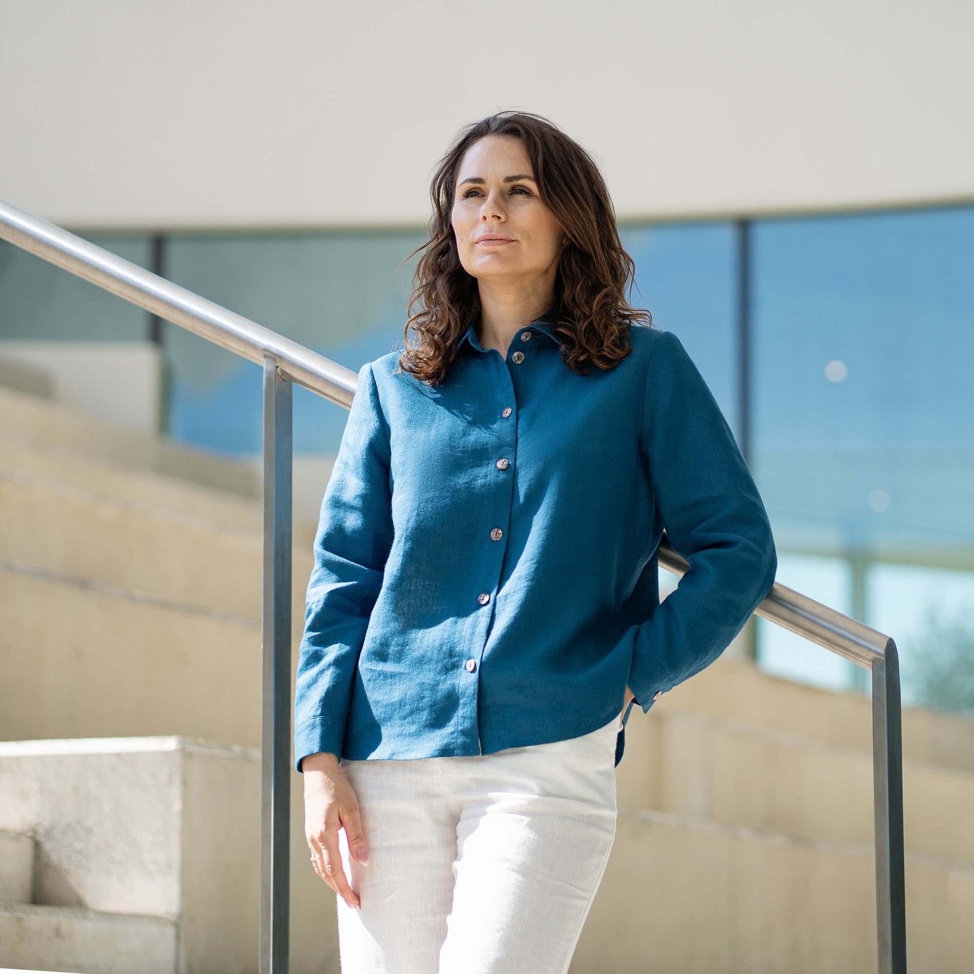 A woman with brown hair, wearing a cobalt blue button-front shirt and wide-leg white trousers, stands outdoors against a modern concrete structure.
