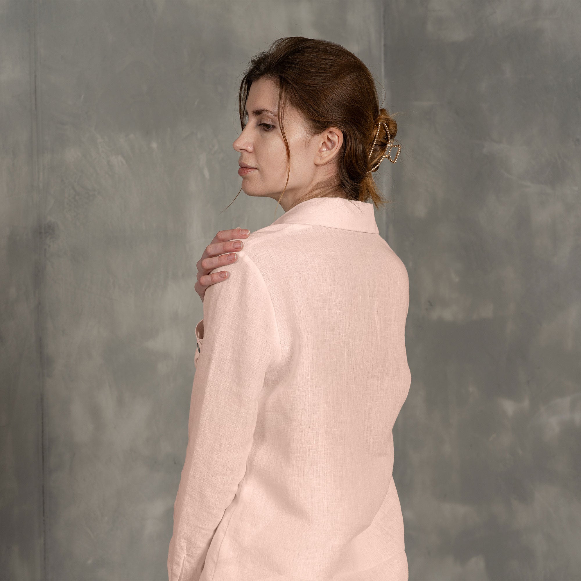 The model is wearing the REBECA classic linen shirt in dusty pink color. She is resting her cheek on her palm, with the other hand in the pants' pocket. The highest button near the collar is undone. It features a regular fit and works well with other officewear clothing pieces.