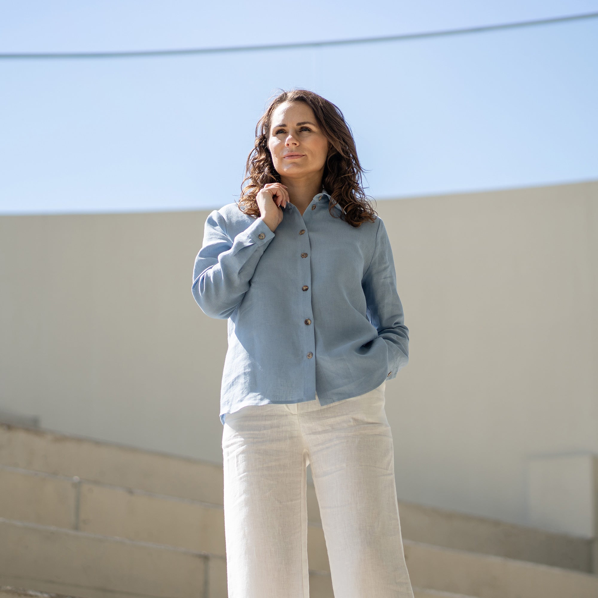 A woman with brown hair, wearing a light blue button-front shirt and wide-leg white trousers, stands outdoors against a modern concrete structure.

