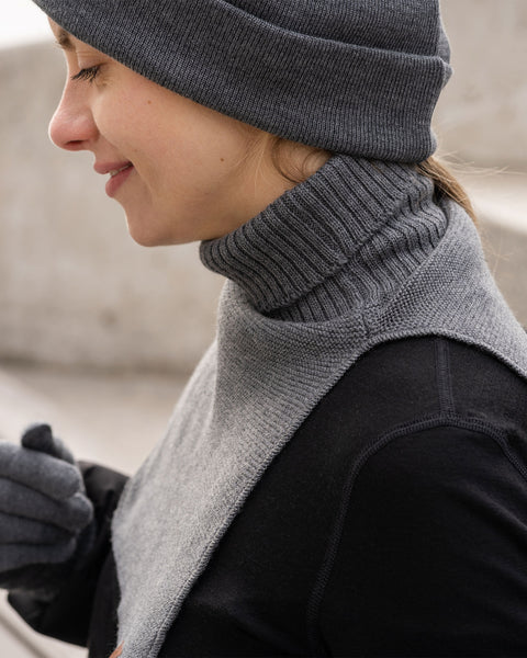 A close-up profile shot of a smiling woman facing left. She is wearing a dark grey knit beanie, a matching dark grey knit neck warmer with a ribbed turtleneck collar and flat bib, and dark grey knit gloves. She is wearing a black long-sleeve shirt underneath, with a dark jacket partially visible near her shoulder. The background consists of blurred light grey concrete steps.