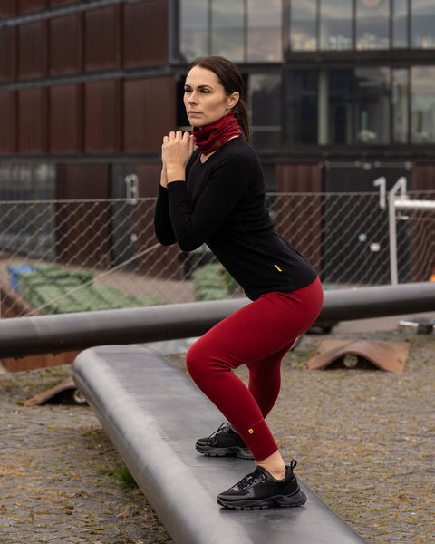 A woman outside in a city, wearing the menique unisex neck gaiter in royal cherry. She is doing leg stretching exercises on a metal object. She also wears a black merino long sleeve top.