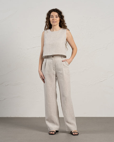 A woman with long, dark curly hair stands facing forward, modeling menique natural linen two-piece outfit. The outfit includes a sleeveless cropped top with a round neckline and high-waisted, wide-legged trousers with pleats at the front. She has one hand in her pocket and is wearing simple black open-toe sandals. The background is a textured light beige wall and a light gray floor.