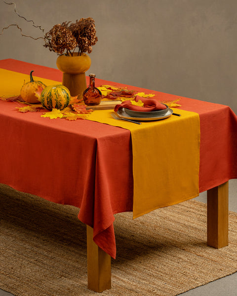 The menique linen tablecloth in cinnamon red color, draping over a wooden table, with a vase on top of it.