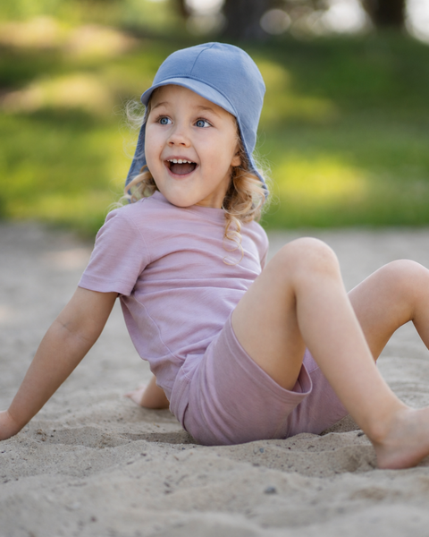 A happy toddler sitting in the sand looking off-camera with an excited expression, wearing a dusty pink matching short-sleeve top and shorts set along with a dusty blue sun cap featuring a neck cover.