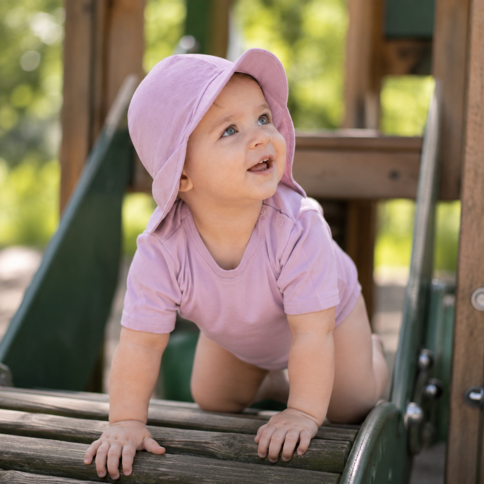 A close-up of a happy baby looking upwards with a little drool around their mouth, wearing a dusty pink short-sleeve top with shoulder snaps and a matching dusty pink sun cap featuring a front brim and neck cover.