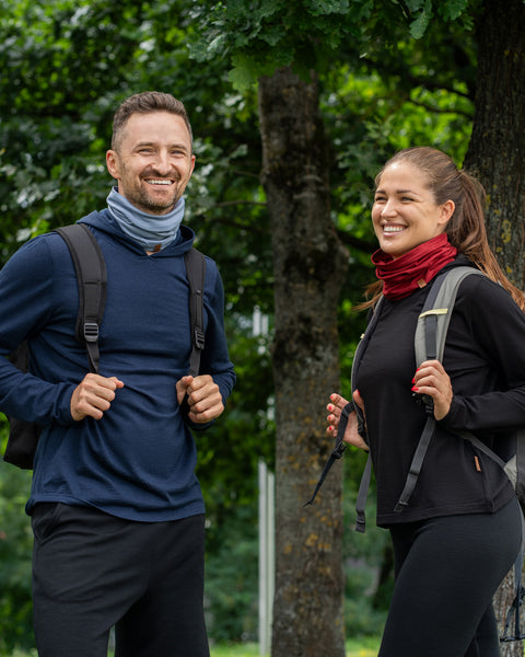A smiling man and woman standing outdoors in a park-like setting. The man on the left is wearing a dark blue long-sleeved hoodie, a backpack, and a light dusty blue neck gaiter scrunched around his neck. The woman on the right is wearing a black long-sleeved hoodie, a backpack, and a deep red neck gaiter scrunched around her neck.