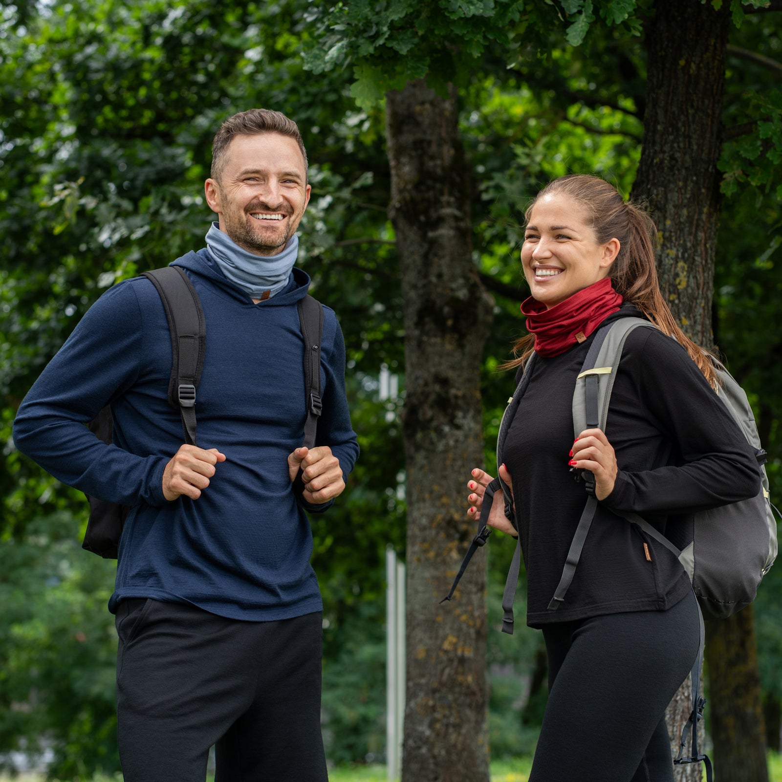 A smiling man and woman standing outdoors in a park-like setting. The man on the left is wearing a dark blue long-sleeved hoodie, a backpack, and a light dusty blue neck gaiter scrunched around his neck. The woman on the right is wearing a black long-sleeved hoodie, a backpack, and a deep red neck gaiter scrunched around her neck.