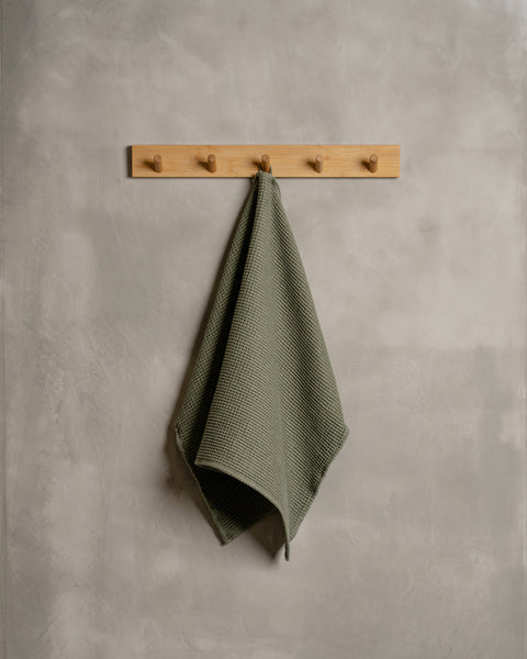 A gray wall with a wooden hanger. In the middle of it hangs a waffle hand washcloth in stone green color.
