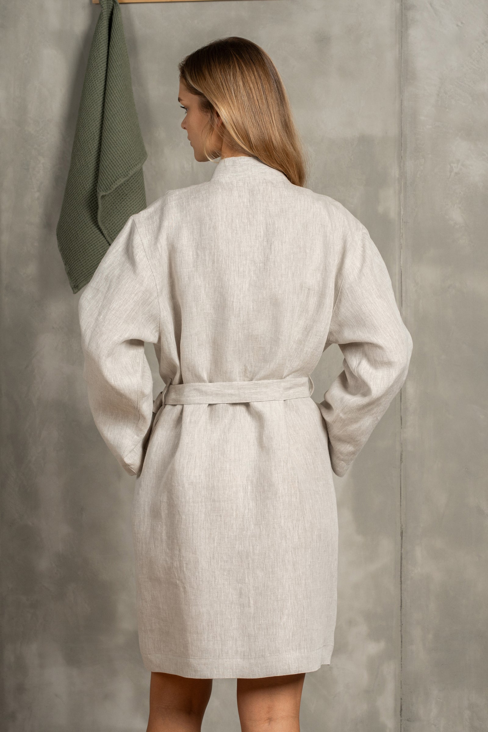 The menique women's linen bathrobe in natural color. The woman is standing in front of a gray wall with a wooden hanger on it. There is a linen waffle towel on the hanger. She has in the pocket, the other put lightly on her stomach.