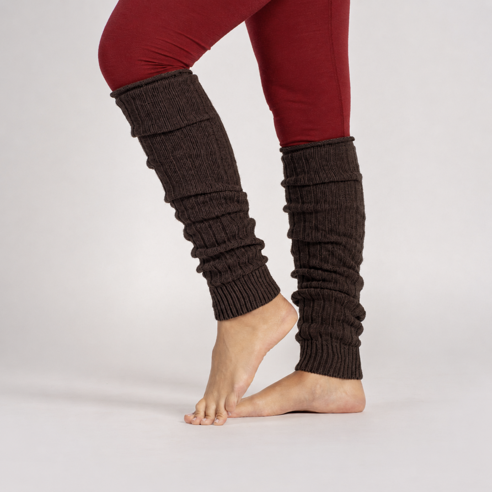 A view of a sitting woman barefoot, wearing brown, ribbed-knit leg warmers scrunched down against a solid light gray background.