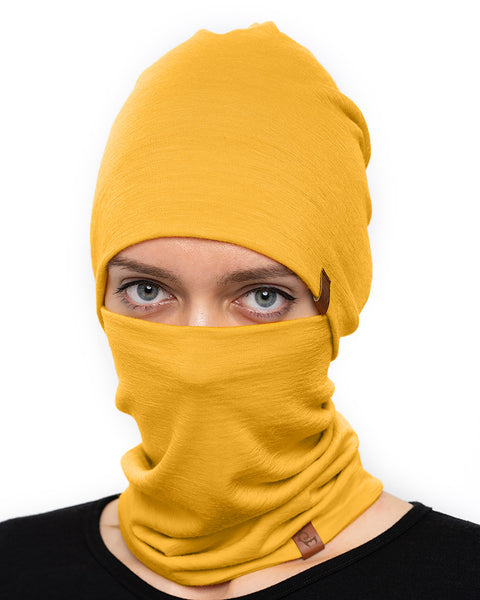 A person with fair skin and light-colored eyes is looking directly at the camera, wearing a spicy yellow menique beanie and a matching neck gaiter that covers their mouth and nose. They are wearing a black garment, and the background is plain white.