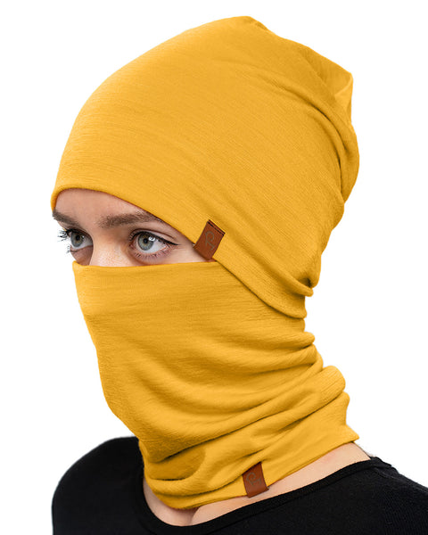 A fair-skinned individual with light-colored eyes is wearing a spicy yellow merino wool beanie that covers their head and a matching neck gaiter pulled up over their mouth and nose, leaving only their eyes visible. They are wearing a black top, and the background is plain white.