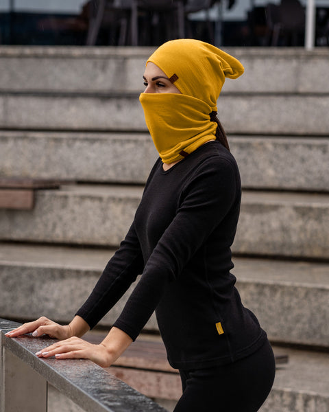 A person with long, dark hair pulled back in a ponytail is standing outdoors, leaning slightly forward with their hands resting on a metal railing. They are wearing a spicy yellow beanie and a matching neck gaiter that covers their mouth and nose, along with a black long-sleeved shirt and black leggings. The background shows a set of wide, light-colored steps leading upwards.