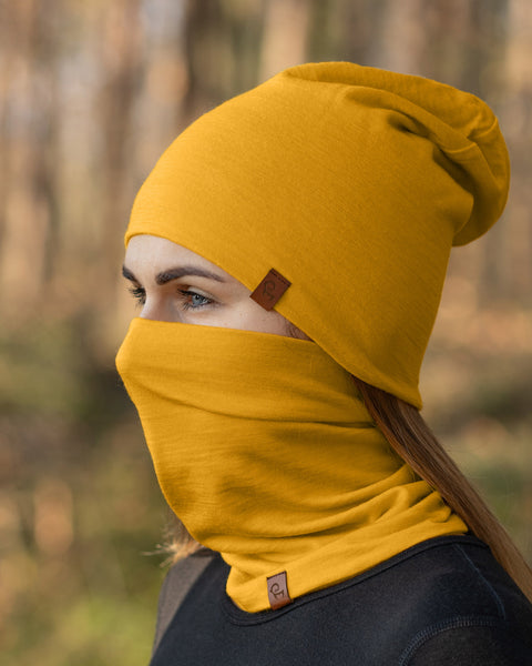 A person with long, light brown hair and fair skin is shown in a profile view, wearing a spicy yellow beanie and a matching neck gaiter that covers their mouth and nose, leaving their light-colored eyes visible. They are wearing a dark-colored garment, and the blurred background suggests an outdoor setting with trees.
