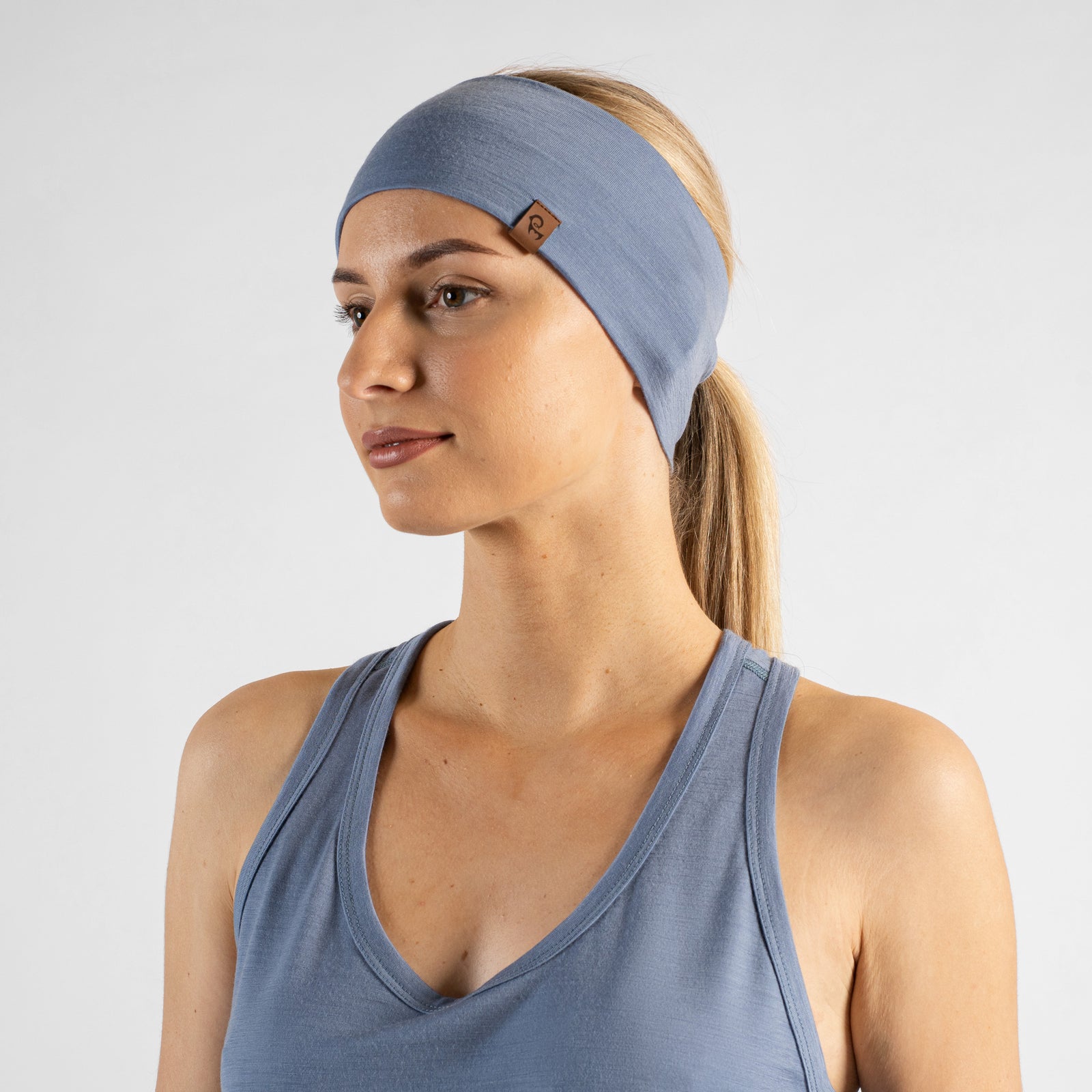 A studio portrait of a blonde woman looking directly at the camera with her body angled slightly away. She is wearing a dusty blue headband over her pulled-back hair, featuring a small brown leather logo patch. She is wearing a matching dusty blue sleeveless top against a plain light grey background.