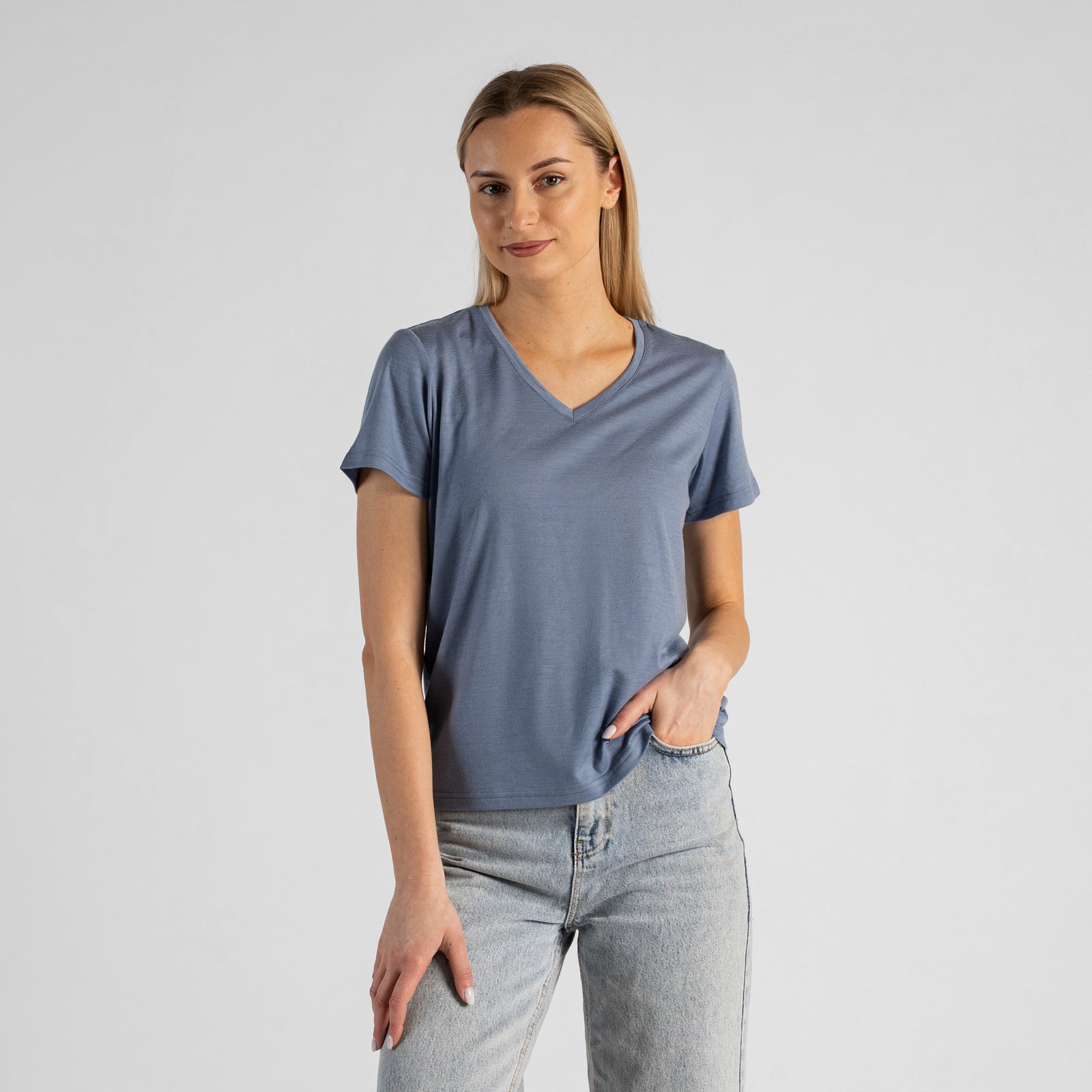 A studio portrait of a blonde woman sitting on a stool and looking directly at the camera. She is leaning back slightly, with her hands resting on her lap. She is wearing a regular fit, dusty blue short-sleeved v-neck t-shirt and light-wash blue jeans against a plain light grey background.