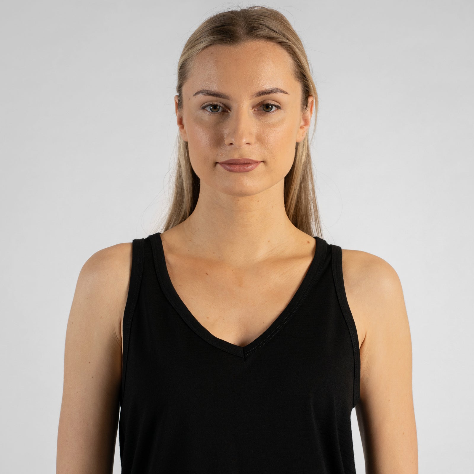 A studio portrait of a blonde woman standing in a three-quarter profile and looking directly at the camera. She is wearing a black sleeveless V-neck top and light wash denim jeans, with one hand resting on her hip against a plain light grey background.