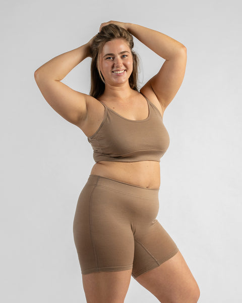 Front three-quarter view of a model holding her hair up with both hands, wearing a beige sports bra and matching beige biker shorts.