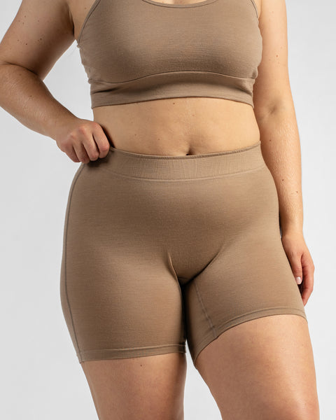 Front view of a model with one hand on her hip, wearing a beige sports bra featuring a black elastic band with text and matching beige biker shorts.