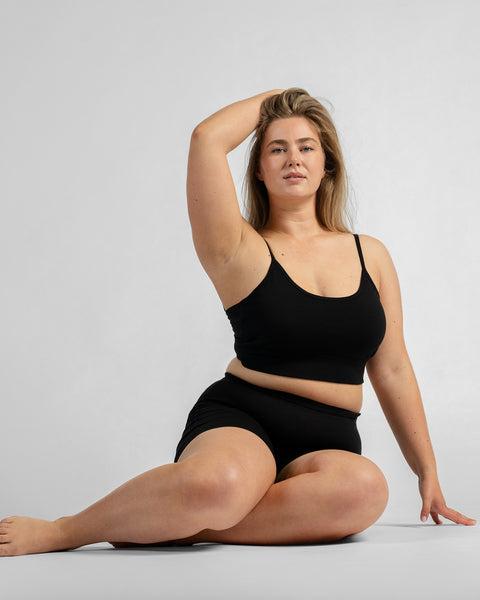 Front full-body view of a model sitting on the floor with one hand behind her head and the other resting on her leg, wearing a black sports bra with thin straps and black biker shorts.
