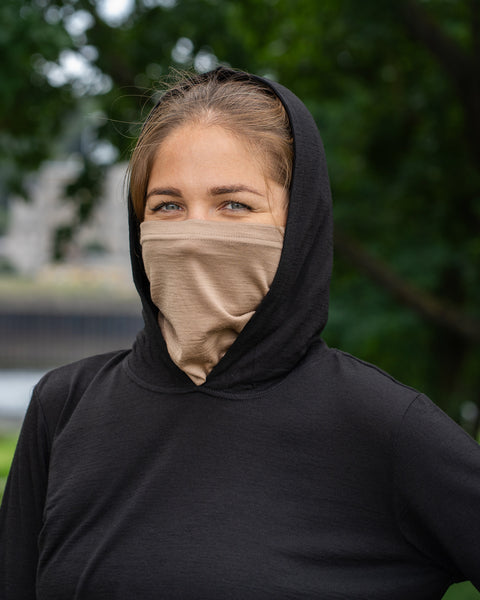 A woman outdoors wearing a black long-sleeved top with the hood pulled up over her head. She is wearing a beige neck gaiter pulled up high to cover her nose and mouth, looking directly at the camera against a backdrop of blurred green trees.