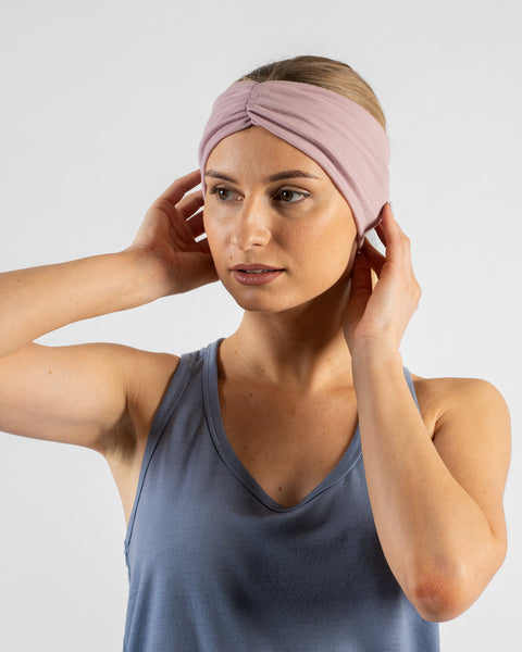A studio portrait of a blonde woman looking slightly off-camera to her left against a plain light grey background. She is wearing a dusty blue sleeveless top and a dusty pink ribbon headband, with her hands gently adjusting the headband near her ears.