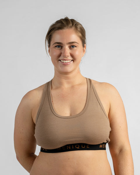 Front view of a smiling model with her arms behind her back, wearing a beige sports bra featuring a black elastic band with text.
