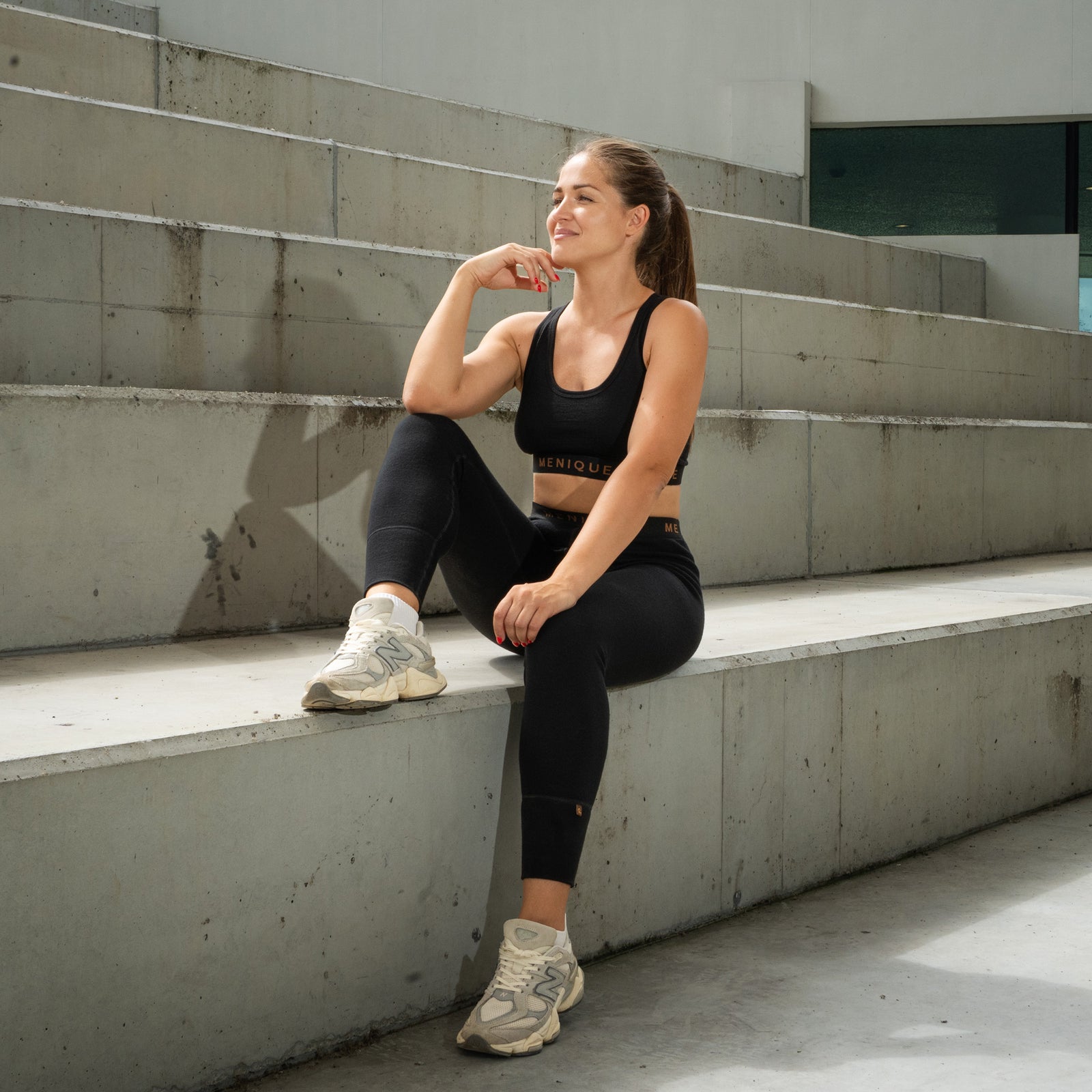 A woman stretching her legs on concrete outdoor stairs by elevating one foot on a higher step, wearing a black sports bra, black leggings, and white sneakers.