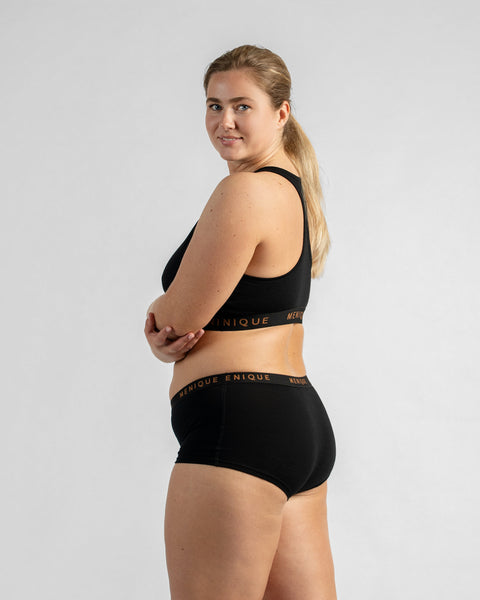 Side view of a model with her arms crossed, looking at the camera, wearing a black sports bra and matching boy-short underwear, both featuring a black elastic band.