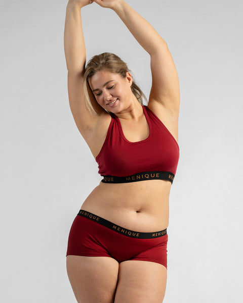 Front view of a model with her arms resting behind her head, wearing a burgundy sports bra and matching boy-short underwear, both featuring a black elastic band.