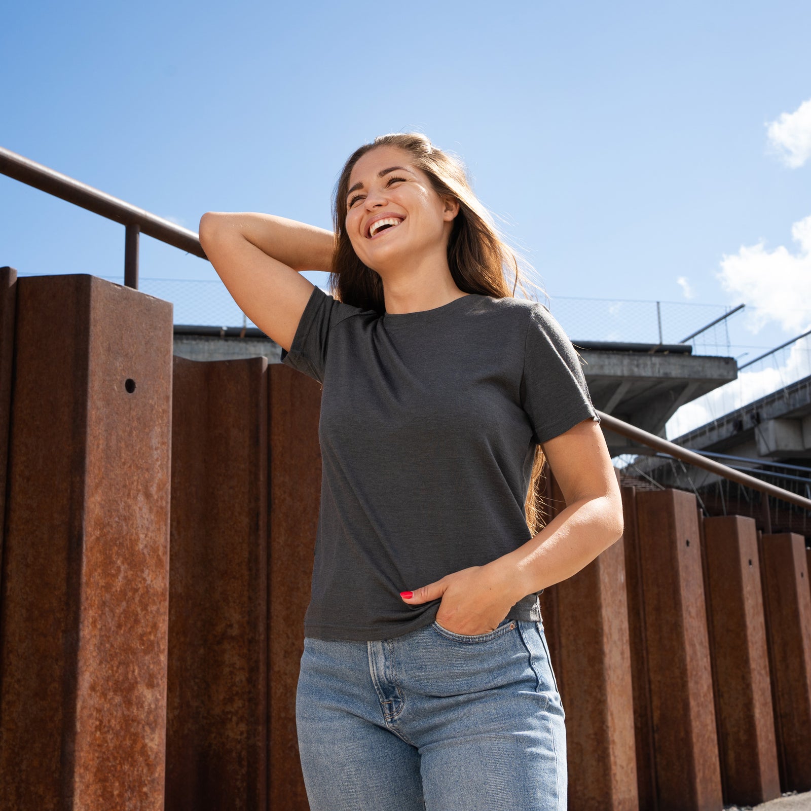 A woman sitting on a paved surface with white parking lines, looking directly up at the camera with a bright smile. She is wearing a charcoal grey short-sleeve crew neck t-shirt and blue jeans.