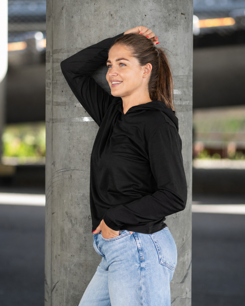 A smiling woman with a ponytail stands outdoors, leaning slightly against a concrete pillar beneath an overpass. She wears a black long-sleeve hoodie and light blue jeans, casually resting one hand near her head and the other in her pocket.