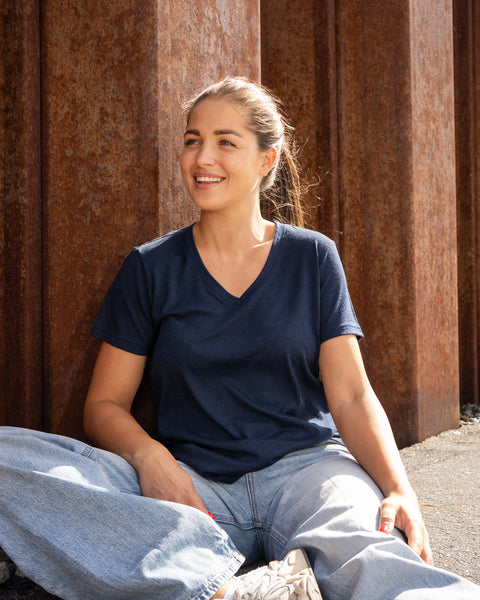 A smiling woman sitting cross-legged on the ground in front of a rust-colored wall, dressed in a dark blue v-neck t-shirt and loose blue jeans.