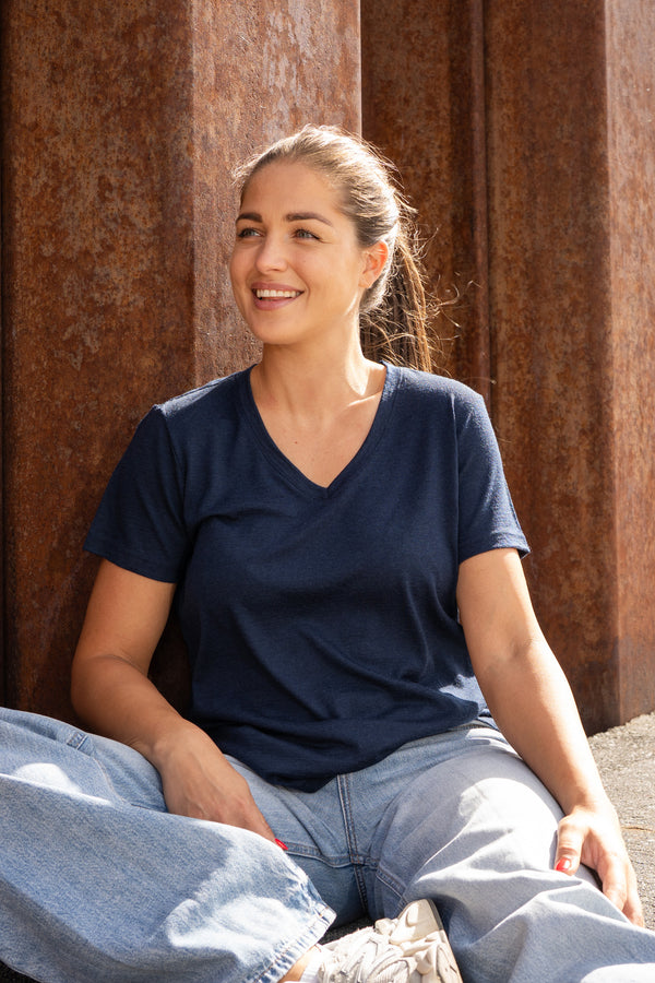 A smiling woman sitting cross-legged on the ground in front of a rust-colored wall, dressed in a dark blue v-neck t-shirt and loose blue jeans.