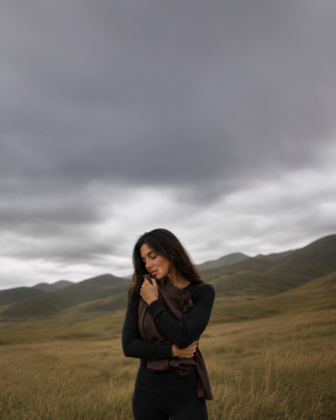 A woman in a black base layer set standing in a hilly, overcast landscape with her eyes closed, clutching a folded brown garment close to her chest.
