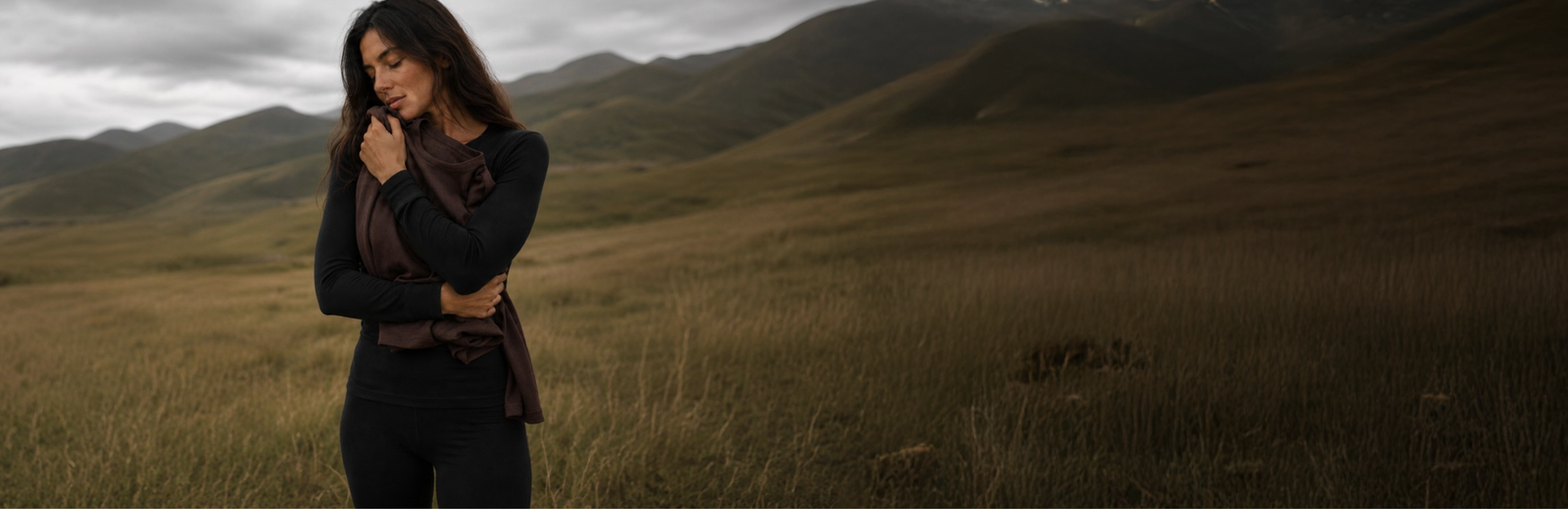 A woman in a black base layer set standing in a hilly, overcast landscape with her eyes closed, clutching a folded brown garment close to her chest.