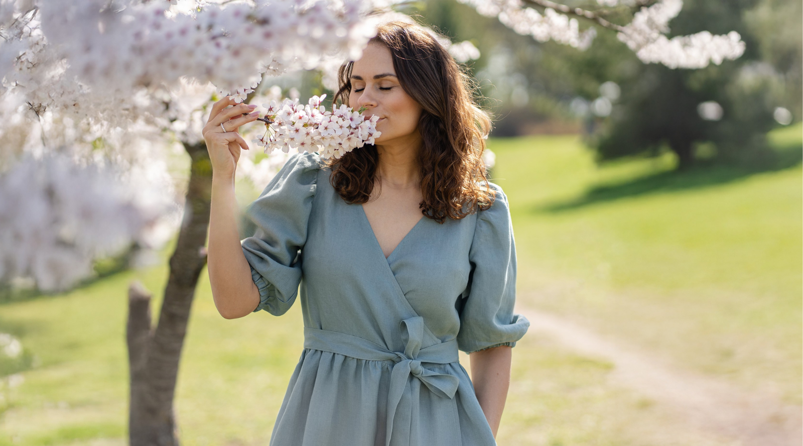Woman wearing light green linen wrap dress smelling spring blossom tree outdoors, breathable women’s linen dress designed for warm weather, natural fabric clothing for spring and summer.