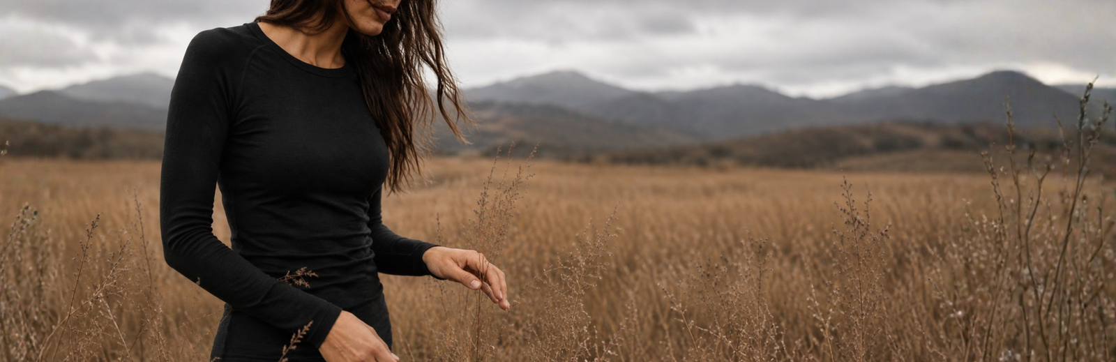 A woman wearing a fitted black long-sleeved top stands in a vast field of dry, golden grass. She gently touches a tall plant stalk with her hand, framed by distant mountains and a cloudy, overcast sky.