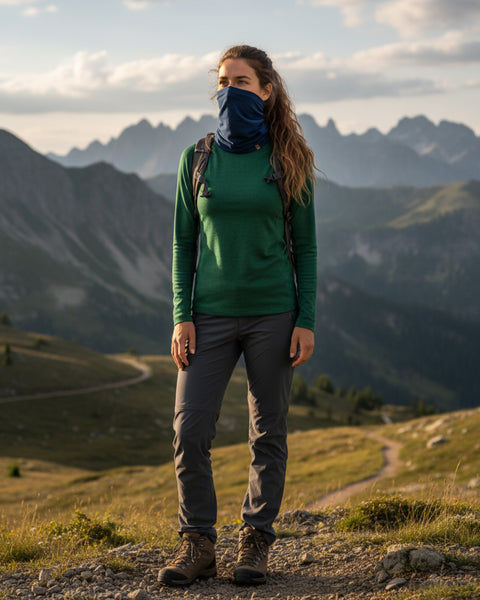 Hiker wearing a denim-blue merino neck gaiter and green top standing on a ridge at sunset, mountains and trails behind.
