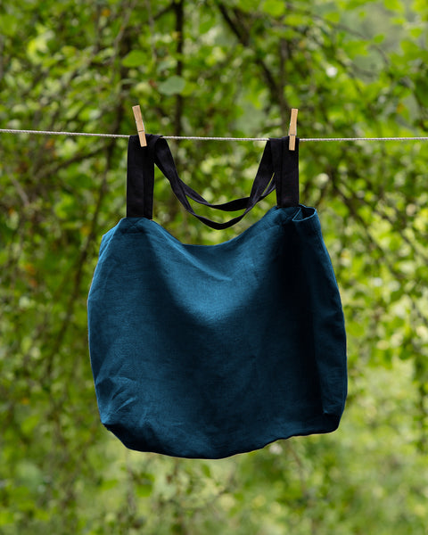 The menique linen tote bag in cobalt blue color with black handles hung on a clothesline outside.
