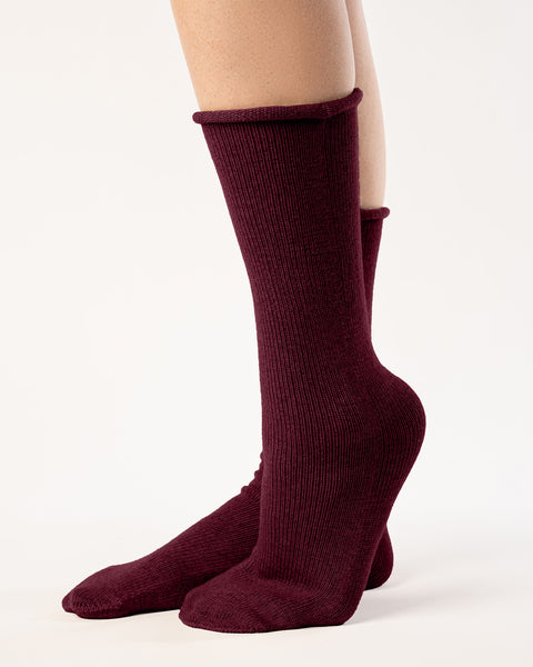 A side view shows a person from the mid-thigh down, wearing ribbed, menique red socks. The socks reach the mid-calf, and the person&#39;s feet are positioned with one slightly in front of the other, heel raised. They stand against a plain white background with even lighting that emphasizes the texture of the socks.