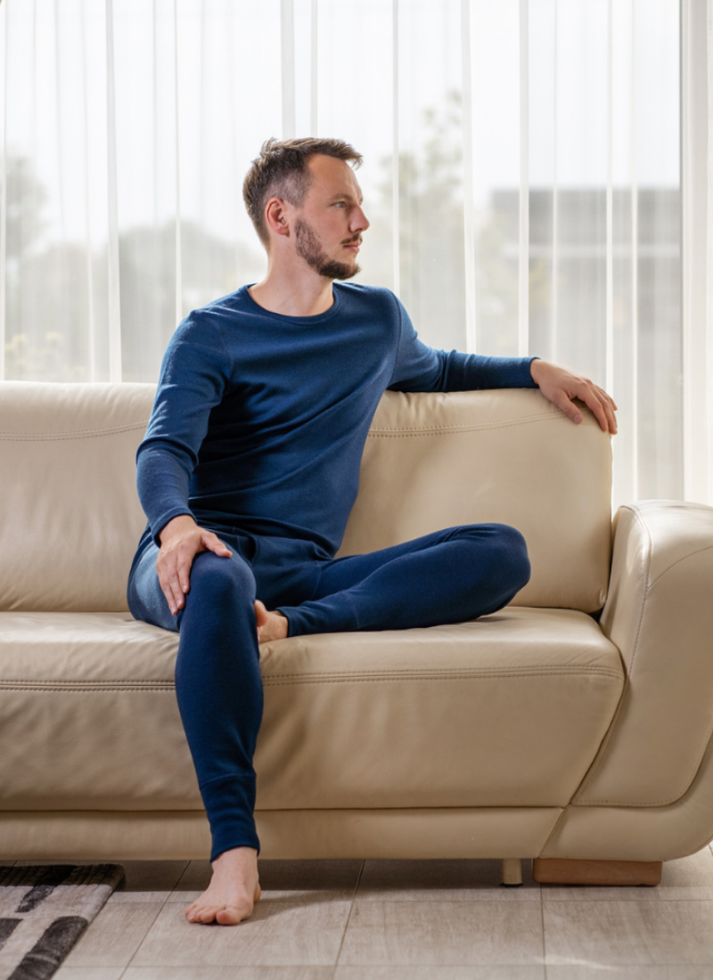 A barefoot man wearing a navy blue long-sleeved base layer set sits on a cream leather sofa indoors. He rests one arm on the back of the sofa and looks off to the side toward a window with sheer white curtains.