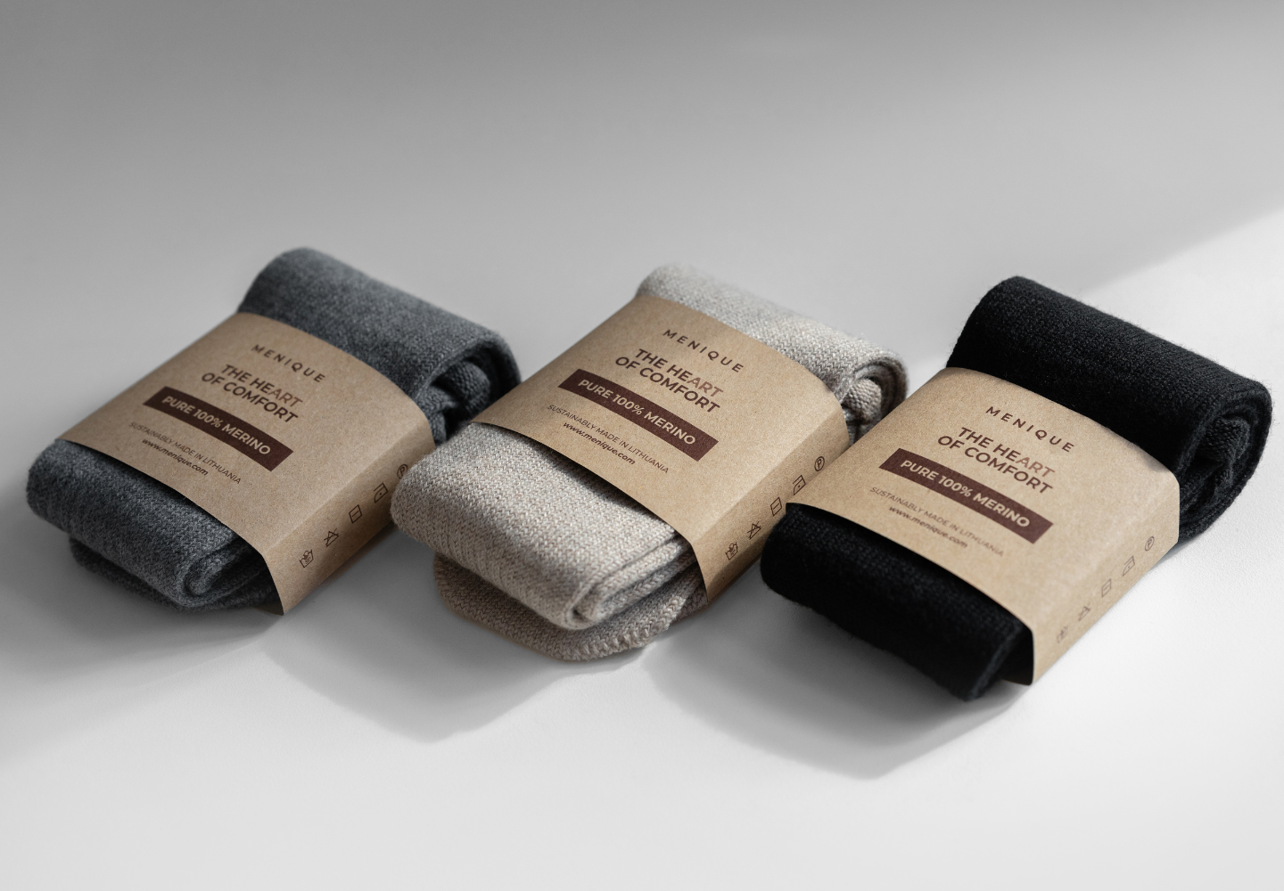 Three pairs of folded merino wool socks from menique in dark grey, beige, and black, each wrapped in eco-friendly kraft paper packaging labeled 'The Heart of Comfort – Pure 100% Merino'.